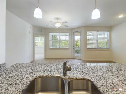 Apartments in Bluffton A modern kitchen with a granite countertop, double sink, and view of an open-concept living area featuring large windows, a ceiling fan, and a balcony.