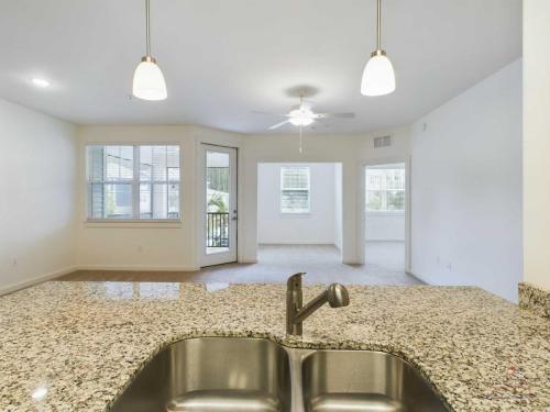 Apartments in Bluffton A modern kitchen with a granite countertop island and a stainless steel double sink overlooks an open-concept living area with large windows, a ceiling fan, and an adjacent room with carpet flooring.