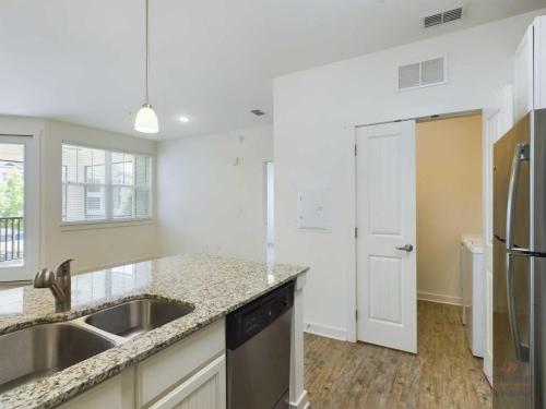 Apartments in Bluffton A modern kitchen with granite countertops, stainless steel appliances, and a double sink. In the background is an open window, and to the right, a door to a laundry room with a washer and dryer.