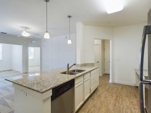 Apartments in Bluffton A modern kitchen with granite countertops, stainless steel appliances, pendant lighting, and a view of the adjoining living area. The space features a mix of wooden flooring and carpeting.