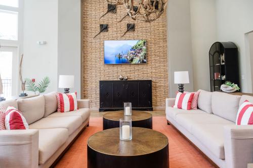 Apartments in Bluffton A modern living room features two beige sofas with red and white pillows, wooden coffee tables with candles, a large wall-mounted TV, and a brick accent wall.