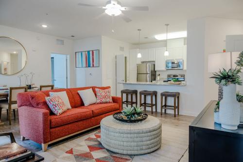 Apartments in Bluffton A modern living room with a red sofa, round wicker coffee table, and a wall mirror. The open kitchen has stainless steel appliances, bar stools at the counter, and a dining area with a wooden table.