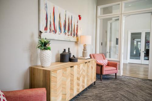 Apartments in Bluffton A modern lobby features a wooden sideboard with decorative items, two coral armchairs with patterned cushions, and a colorful abstract painting on the wall.