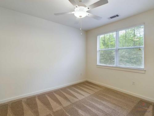 Apartments in Bluffton A room with beige carpet, white walls, a ceiling fan, and a window with blinds. The floor is vacuumed with visible lines.