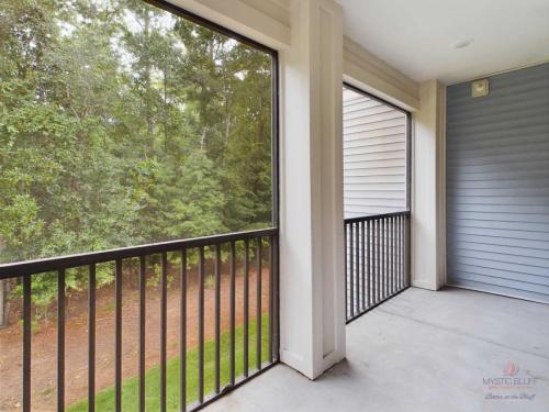 Apartments in Bluffton A screened-in balcony with a black railing overlooks a grassy area and trees. The space features beige walls and a blue exterior wall. A white ceiling light is visible.