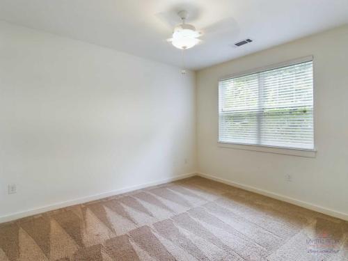 Apartments in Bluffton A simple, empty room with white walls, tan carpet with a pattern, a ceiling fan with a light, and a window with horizontal blinds.