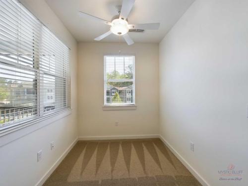 Apartments in Bluffton A small, empty room with beige carpet and white walls, featuring a ceiling fan with light and two windows letting in natural daylight.