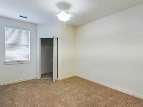 Apartments in Bluffton A small, empty room with beige carpet, white walls, a ceiling fan, a window with blinds, and an open closet door.