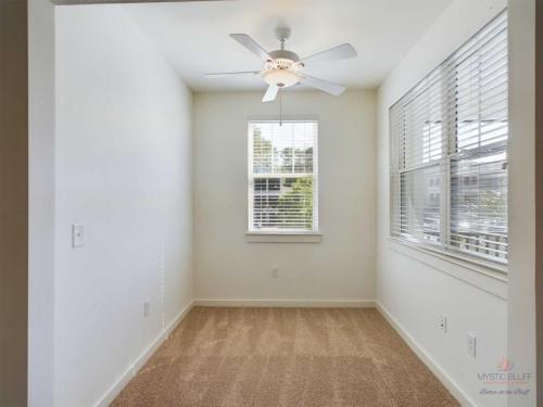 Apartments in Bluffton A small, empty room with beige carpet, white walls, a ceiling fan with a light, and windows with blinds. There is natural light coming through the windows.