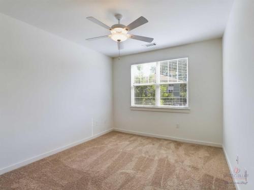 Apartments in Bluffton A small empty room with beige carpet, white walls, a ceiling fan with light, and a window with blinds letting in natural light.