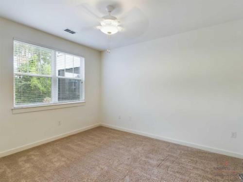 Apartments in Bluffton A small, empty room with white walls, a brown carpet, a ceiling fan with a light, and a window with closed blinds that overlooks greenery.