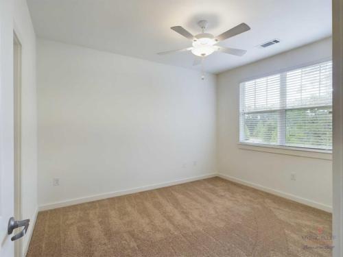 Apartments in Bluffton A small empty room with white walls, a ceiling fan with a light, a window with blinds, and beige carpet flooring.