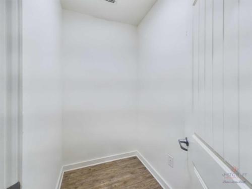 Apartments in Bluffton A small, empty room with white walls, a wooden floor, and a white door slightly ajar. A vent is visible on the ceiling.