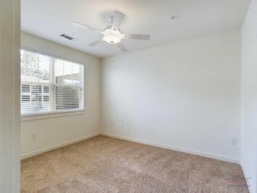 Apartments in Bluffton A small, empty room with white walls, brown carpet, a ceiling fan with a light, and a window with blinds.