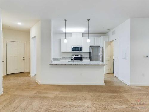 Apartments in Bluffton A small kitchen with white cabinets, stainless steel appliances, and granite countertops. Two pendant lights hang above the kitchen island. The space has brown carpet and white walls.