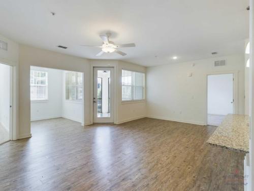 Apartments in Bluffton A spacious, empty living room with wooden flooring, white walls, a ceiling fan, large windows, and a glass door leading to a patio. A kitchen counter is visible on the right side of the image.