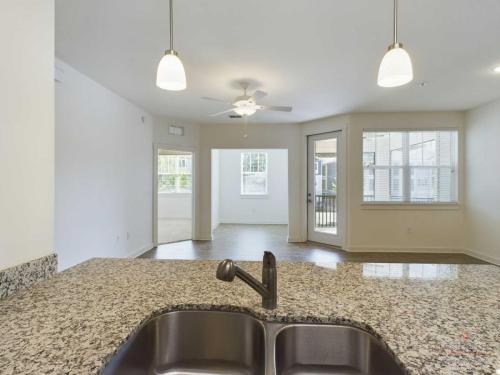 Apartments in Bluffton A spacious modern kitchen with granite countertops and stainless steel sink overlooks an open-plan living area with large windows, ceiling fan, and access to a balcony.