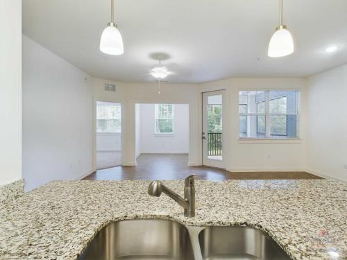 Apartments in Bluffton A view from a kitchen with a granite countertop and double sink looking into an open-plan living room with large windows, a ceiling fan, and a door leading to an outdoor balcony.