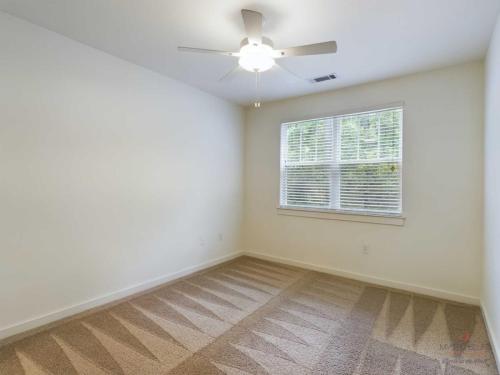 Apartments in Bluffton An empty room with beige carpet, white walls, a window with closed blinds, and a ceiling fan with light.