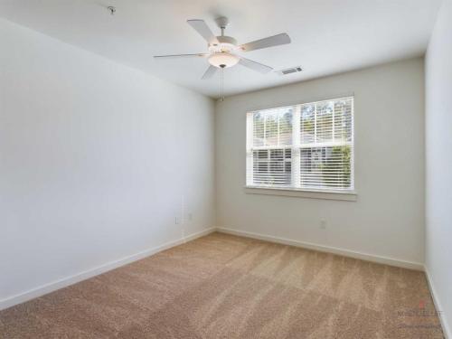 Apartments in Bluffton An empty room with white walls, a ceiling fan, beige carpet, and a window with blinds letting in natural light.