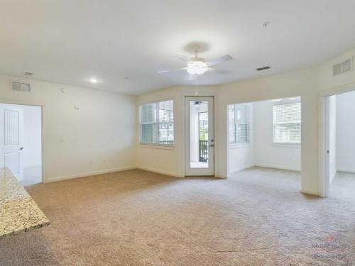 Apartments in Bluffton Bright living room with beige carpet, ceiling fan, and large windows. An open doorway leads to a balcony. Two adjoining rooms are visible, one with a door. A granite counter is partially visible.