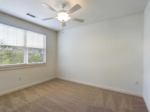 Apartments in Bluffton Empty room with beige carpet, white walls, a large window with blinds, and a ceiling fan with a light fixture.