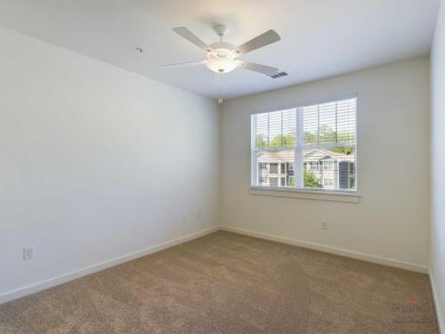 Apartments in Bluffton Empty room with beige carpet, white walls, ceiling fan with light, and a window with blinds. One small light switch and two electrical outlets are on the walls.