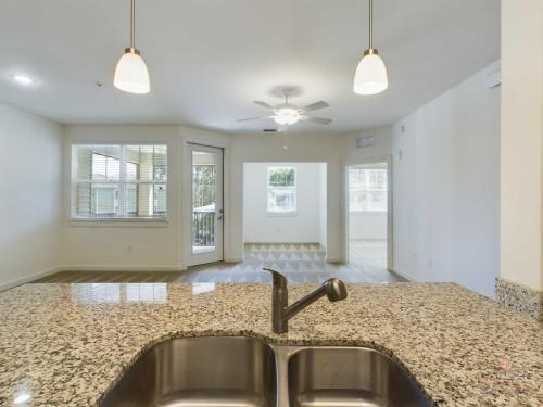 Apartments in Bluffton Granite kitchen countertop with stainless steel sink and faucet, pendant lights above, and an open floor plan leading to a living area with large windows and ceiling fan.