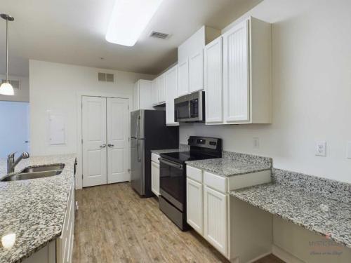 Apartments in Bluffton Modern kitchen with granite countertops, white cabinetry, stainless steel appliances, and a double sink. Wood flooring throughout and a pantry with double doors in the background.