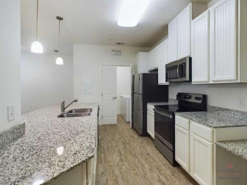 Apartments in Bluffton Modern kitchen with white cabinets, granite countertops, stainless steel appliances, and wood-style flooring. There is a door at the end of the kitchen and recessed lighting on the ceiling.