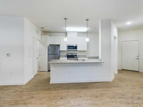 Apartments in Bluffton Modern kitchen with white cabinets, stainless steel appliances, granite countertops, and pendant lights. Wood flooring extends throughout the open space, with a front door visible to the right.