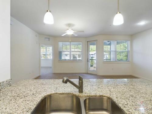 Apartments in Bluffton Modern open-plan apartment with a granite kitchen counter, stainless steel sink, pendant lights, and ceiling fan. Large windows and French doors leading to a balcony offer ample natural light.