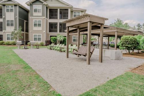 Apartments in Bluffton Outdoor area with wooden pergola, swing, and several green Adirondack chairs on gravel ground, located in front of multi-story residential buildings with balconies and surrounded by greenery.
