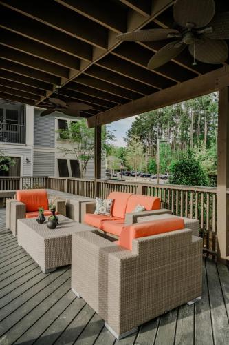 Apartments in Bluffton Outdoor patio with beige wicker furniture, bright orange cushions, and a wooden deck. Ceiling fans are mounted on the overhead wooden beams. Trees and a building are visible in the background.