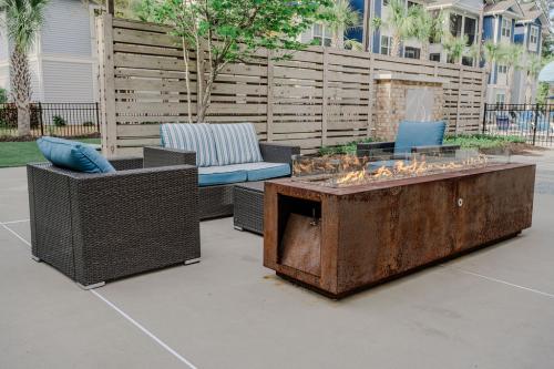 Apartments in Bluffton Outdoor seating area with wicker sofa and armchair featuring blue cushions, centered around a rectangular metal fire pit. Wood fence and apartment buildings are visible in the background.