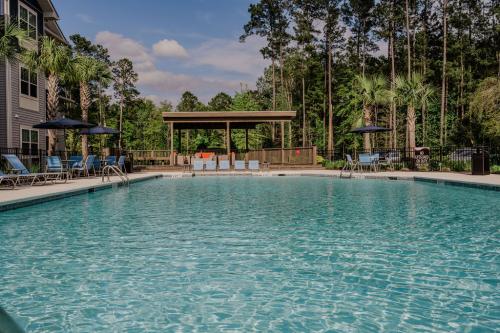 Apartments in Bluffton Outdoor swimming pool with lounge chairs, umbrellas, and a wooden pavilion. Trees and a blue sky can be seen in the background.