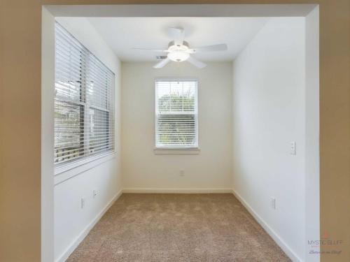 Apartments in Bluffton Small, empty room with beige carpet, off-white walls, and a ceiling fan with a light. Two large windows on the left wall let in natural light.