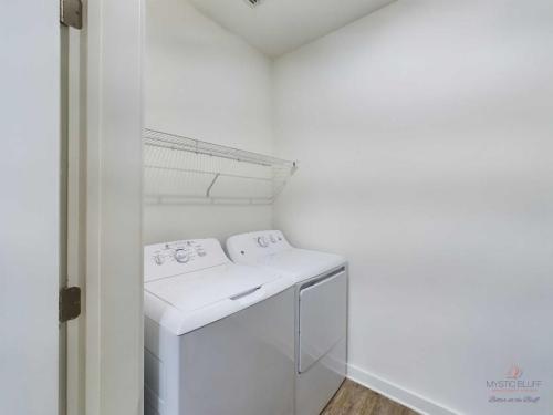 Apartments in Bluffton Small laundry room with a white washing machine and dryer side by side, an overhead wire shelf, and white walls.