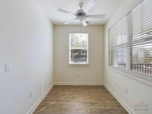 Apartments in Bluffton Small, white-walled room with a wood floor, ceiling fan, and two windows with blinds, letting in natural light.