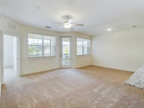 Apartments in Bluffton Spacious room with beige carpet, large windows, a ceiling fan, and a door leading to a balcony. Nearby, there is a partially visible room with an open door.