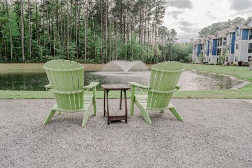 Apartments in Bluffton Two green Adirondack chairs face a pond with a central water fountain, surrounded by trees and apartment buildings in the background. A small wooden table is positioned between the chairs.