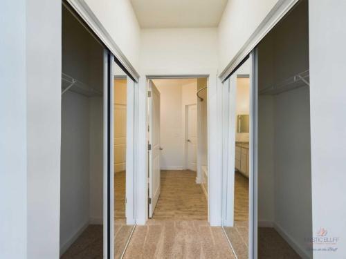 Apartments in Bluffton View of an empty walk-in closet with sliding doors, beige carpet, and white walls, leading to a bathroom with a wooden floor and a vanity on the right.